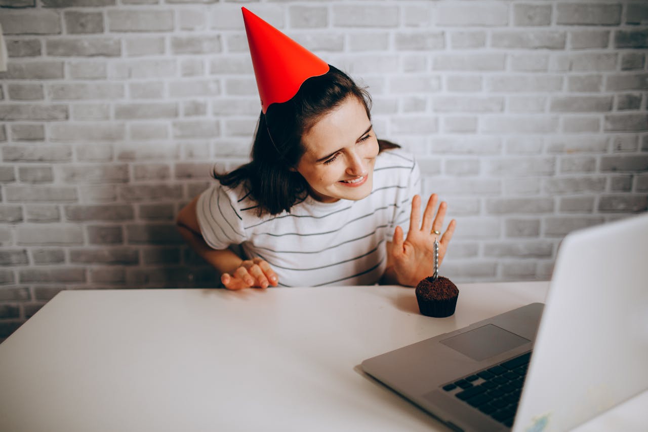 Woman in party hat waving at laptop and cupcake, celebrating virtual birthday.
